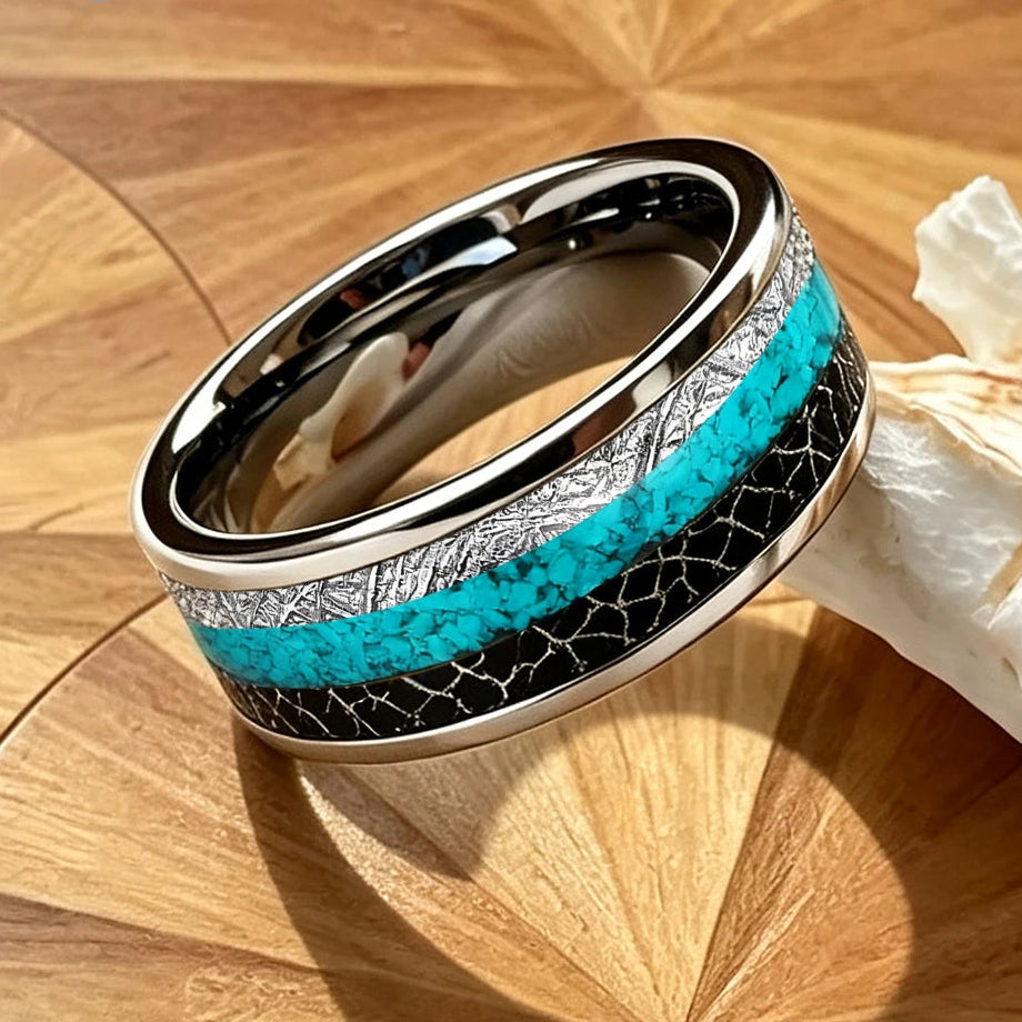 A polished silver-toned band ring with a turquoise inlay and black textured detailing, displayed on a wooden surface next to a white seashell.