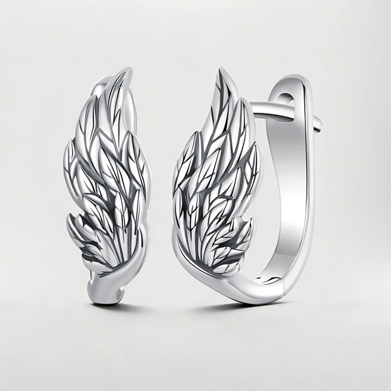 A pair of polished silver huggie hoop earrings featuring detailed wing designs, displayed on a clean light gray background.